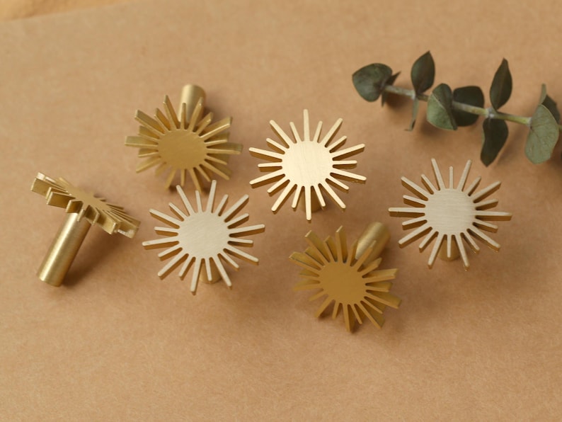 May include: Six gold-toned metal drawer pulls shaped like sunbursts. Each pull has a circular centre and radiating points.