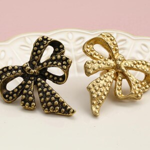 Könnte beinhalten: Zwei schleifenförmige Broschen, eine dunkelbronze und eine goldfarbene, mit einer strukturierten, gepunkteten Oberfläche. Sie liegen auf einem weißen, gewellten Teller vor einem rosa Hintergrund. Diese Broschen sind wahrscheinlich Vintage oder antik.