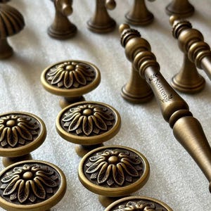 May include: Bronze cabinet hardware with floral and ornate designs. The image shows various knobs and pulls, including oval knobs with a sunflower motif and longer pulls with the text "Notten".