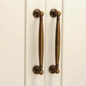 Pu&ograve; includere: Due maniglie per mobili in bronzo antico. Ogni maniglia ha una placca posteriore rotonda decorativa, una lunga maniglia affusolata e un terminale rotondo. Le maniglie sono montate su un'anta bianca.