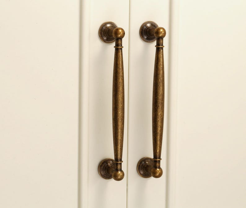 Pu&ograve; includere: Due maniglie per mobili in bronzo antico. Ogni maniglia ha una placca posteriore rotonda decorativa, una lunga maniglia affusolata e un terminale rotondo. Le maniglie sono montate su un'anta bianca.