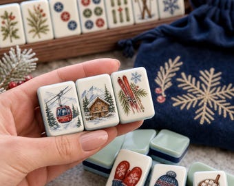 May include: A set of rectangular mahjong tiles featuring winter-themed illustrations including a red cable car, a wooden cabin, skis, red mittens, a knit hat with goggles, and a mug of hot cocoa, stored in a dark blue velvet drawstring bag.