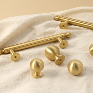 K&ouml;nnte beinhalten: Eine Sammlung goldfarbener Schrankbeschl&auml;ge, darunter Stangengriffe und runde Kn&ouml;pfe. Die Beschl&auml;ge haben eine glatte, metallische Oberfl&auml;che und sind auf einem cremefarbenen Stoff angeordnet. Die Kn&ouml;pfe und Griffe sind f&uuml;r M&ouml;bel und Schrankt&uuml;ren konzipiert.