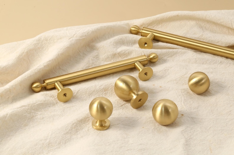 K&ouml;nnte beinhalten: Eine Sammlung goldfarbener Schrankbeschl&auml;ge, darunter Stangengriffe und runde Kn&ouml;pfe. Die Beschl&auml;ge haben eine glatte, metallische Oberfl&auml;che und sind auf einem cremefarbenen Stoff angeordnet. Die Kn&ouml;pfe und Griffe sind f&uuml;r M&ouml;bel und Schrankt&uuml;ren konzipiert.