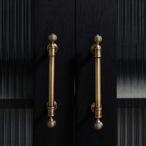 May include: Close-up of a black cabinet door with two antique brass-coloured handles. The handles are vertical, with a decorative, swirled design at the top and bottom. The door has a textured, ribbed glass panel.