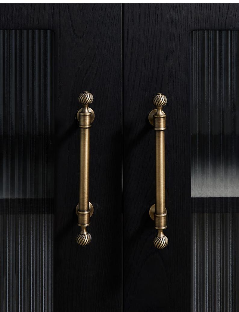 May include: Close-up of a black cabinet door with two antique brass-coloured handles. The handles are vertical, with a decorative, swirled design at the top and bottom. The door has a textured, ribbed glass panel.