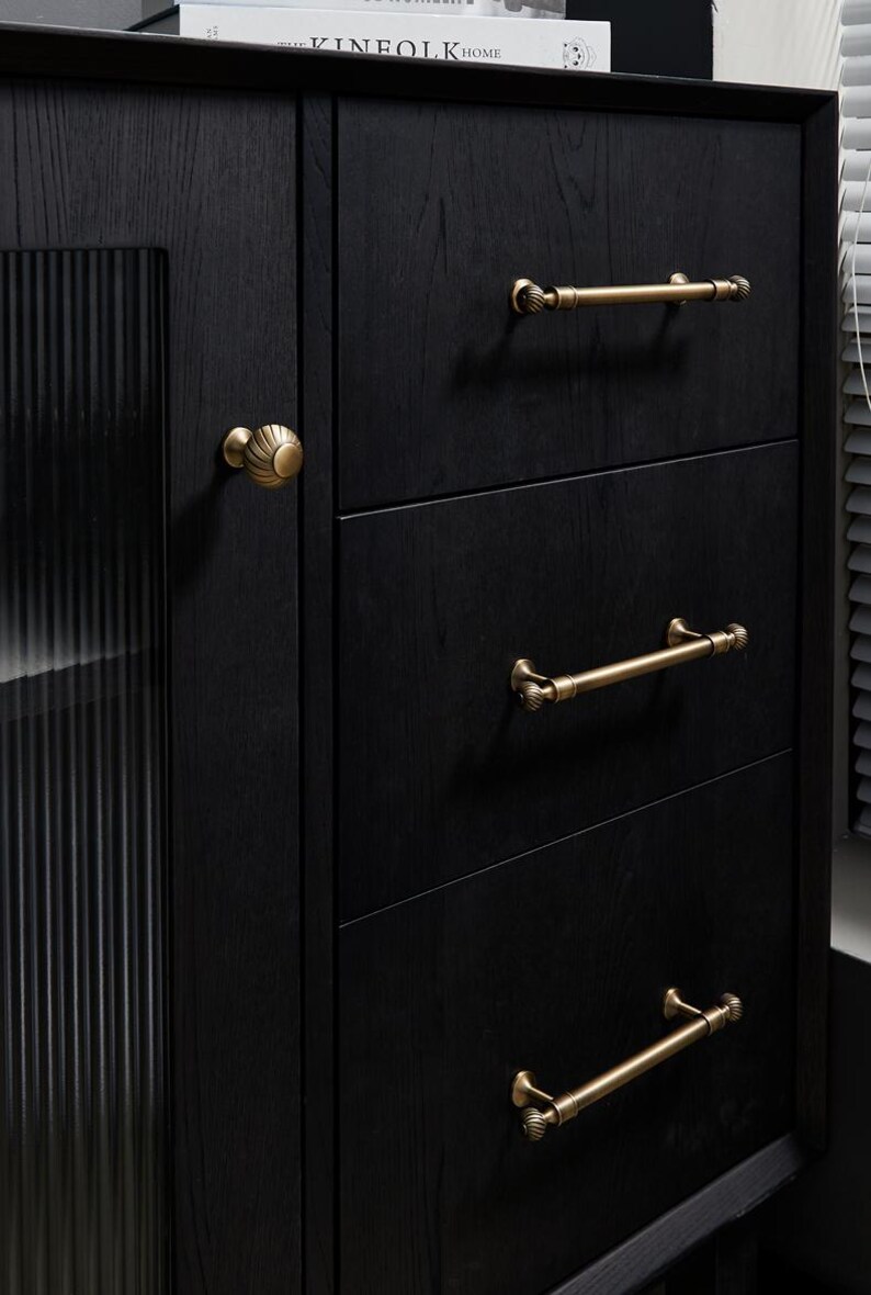 May include: Close-up of a black wooden cabinet with three drawers and a side cabinet. The cabinet has gold-coloured handles and knobs. The side cabinet has a glass door with vertical lines.