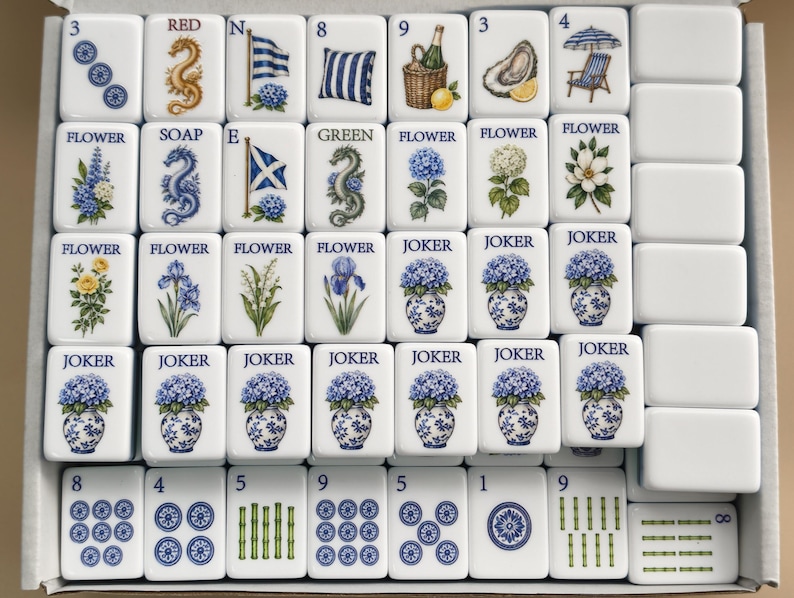 May include: A set of white rectangular Mahjong tiles arranged in a box, featuring blue and green illustrations of flowers, dragons, flags, and various objects. Each tile displays specific text labels like Flower, Joker, Red, Soap, and Green.
