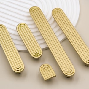 Peut inclure: Quatre poignées de cabinet de couleur or avec un design rainuré. Les poignées sont de forme ovale et ont un bord arrondi. Une poignée est de forme arrondie plus petite.