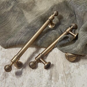 May include: Two antique brass door pulls with a ribbed ball design and circular mounting plates. The pulls are different sizes, with one being longer than the other. They rest on a textured, neutral-toned fabric.