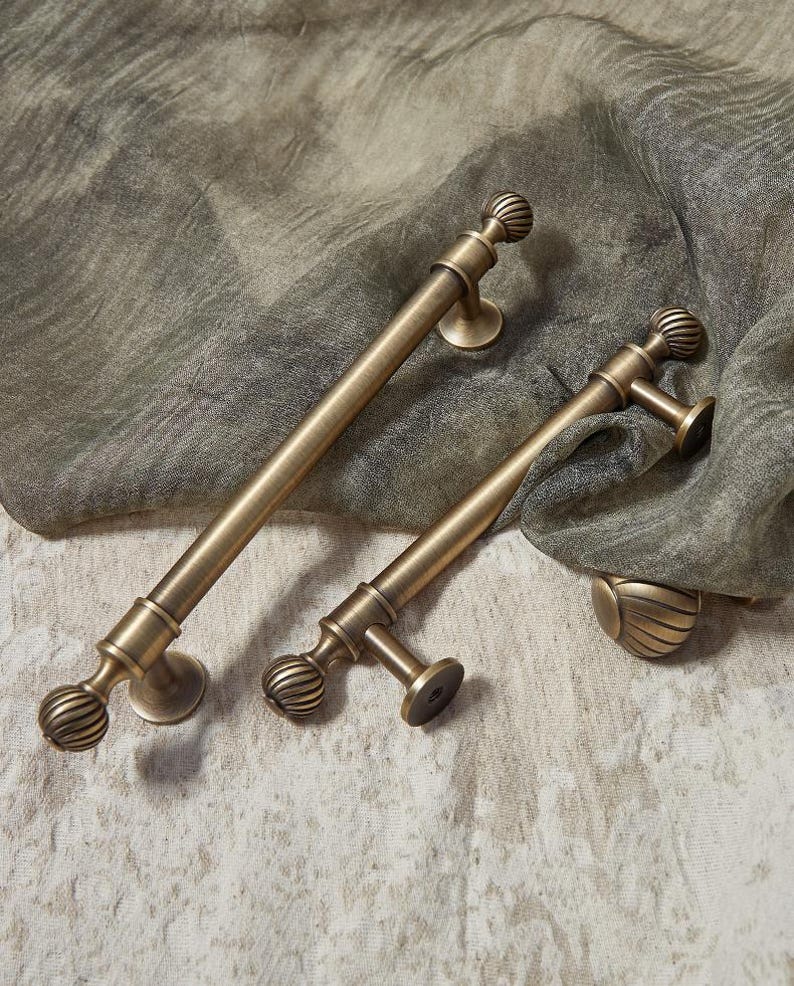 May include: Two antique brass door pulls with a ribbed ball design and circular mounting plates. The pulls are different sizes, with one being longer than the other. They rest on a textured, neutral-toned fabric.