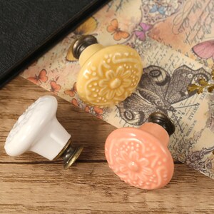 Op de afbeelding: Drie keramische kastknoppen met bloemmotieven. E&eacute;n is wit, &eacute;&eacute;n is lichtgeel en &eacute;&eacute;n is lichtroze.