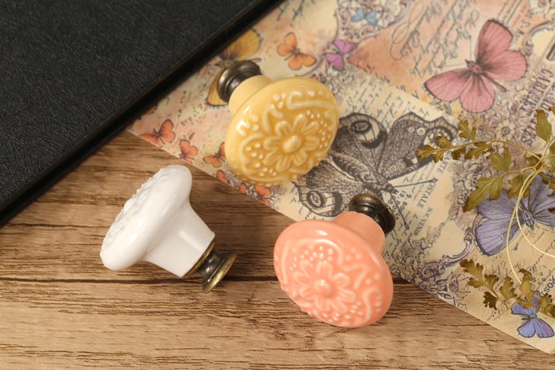 Op de afbeelding: Drie keramische kastknoppen met bloemmotieven. E&eacute;n is wit, &eacute;&eacute;n is lichtgeel en &eacute;&eacute;n is lichtroze.