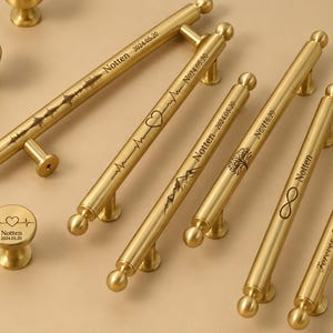 May include: Gold-toned cabinet hardware set featuring various handle styles and matching knobs. The handles are engraved with designs like a mountain range, heartbeat, tree, and infinity symbol, along with the name "Notten" and dates.