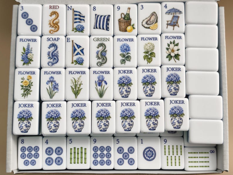 May include: A set of white rectangular Mahjong tiles arranged in a box. The tiles feature blue and green illustrations of flowers, dragons, flags, and various objects, with text labels including Flower, Joker, Red, Green, and Soap, alongside numbered suit tiles.