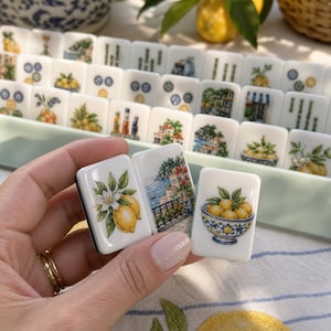 May include: A set of white rectangular mahjong tiles featuring colorful illustrations of lemons, coastal Mediterranean architecture, and bowls of fruit. The tiles are arranged in a light green tray, with three individual tiles held in the foreground.
