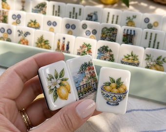 May include: A set of white rectangular mahjong tiles featuring colorful illustrations of lemons, coastal Mediterranean architecture, and bowls of fruit. The tiles are arranged in a light green tray, with three individual tiles held in the foreground.