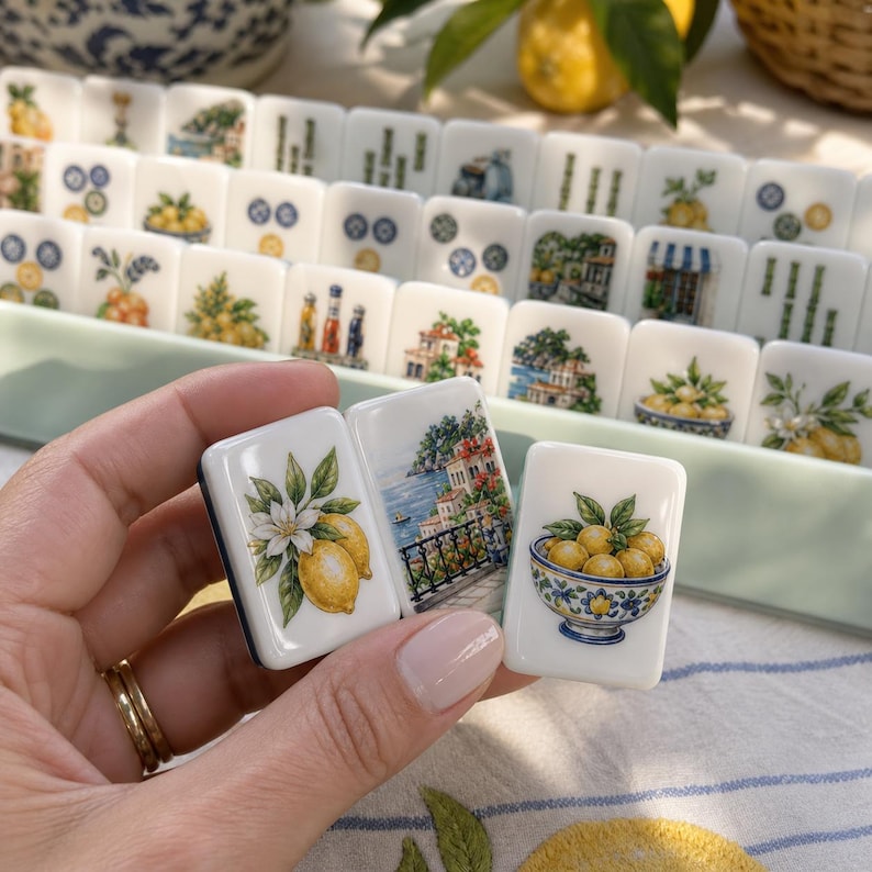 May include: A set of white rectangular mahjong tiles featuring colorful illustrations of lemons, coastal Mediterranean architecture, and bowls of fruit. The tiles are arranged in a light green tray, with three individual tiles held in the foreground.