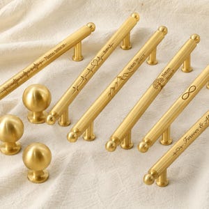 May include: A collection of gold-colored cabinet hardware, including several pull handles and round knobs. The handles feature engraved designs and text, such as a heartbeat, mountains, and the infinity symbol. The hardware is displayed on a cream-colored fabric.