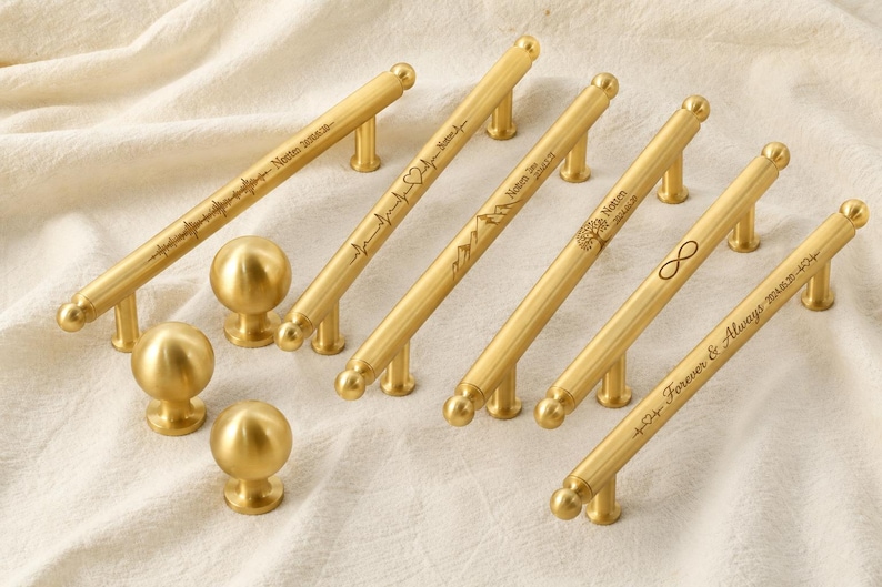 May include: A collection of gold-colored cabinet hardware, including several pull handles and round knobs. The handles feature engraved designs and text, such as a heartbeat, mountains, and the infinity symbol. The hardware is displayed on a cream-colored fabric.