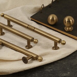 K&ouml;nnte beinhalten: Eine Sammlung antiker Messing-Schrankbeschl&auml;ge, darunter Stangengriffe und ein runder Knauf. Die Beschl&auml;ge haben einen warmen, goldenen Farbton und ein klassisches Design. Das Wort "Notten" ist auf den Stangengriffen aufgedruckt.