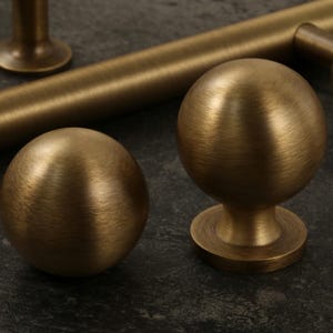 K&ouml;nnte beinhalten: Nahaufnahme von antiken Messing-Schrankbeschl&auml;gen, darunter zwei kugelf&ouml;rmige Kn&ouml;pfe und ein zylindrischer Griff. Die Beschl&auml;ge haben eine geb&uuml;rstete Oberfl&auml;che und sind vor einem dunklen, strukturierten Hintergrund platziert. Die Kn&ouml;pfe haben einen Durchmesser von etwa 5 cm.