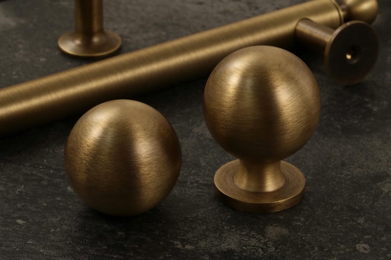 K&ouml;nnte beinhalten: Nahaufnahme von antiken Messing-Schrankbeschl&auml;gen, darunter zwei kugelf&ouml;rmige Kn&ouml;pfe und ein zylindrischer Griff. Die Beschl&auml;ge haben eine geb&uuml;rstete Oberfl&auml;che und sind vor einem dunklen, strukturierten Hintergrund platziert. Die Kn&ouml;pfe haben einen Durchmesser von etwa 5 cm.