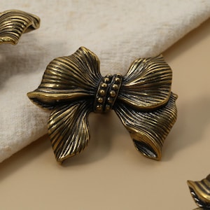 May include: Close-up of antique brass bow-shaped cabinet hardware. The hardware has a detailed, textured design with a vintage aesthetic. The bow is centered with a series of small, round accents. The hardware is on a neutral background.