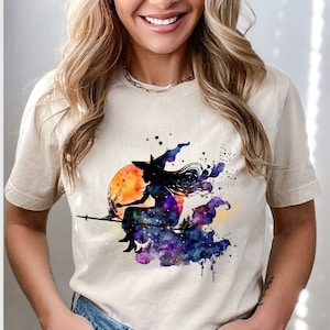 May include: A beige t-shirt with a watercolor design of a witch flying on a broomstick in front of a moon and stars.