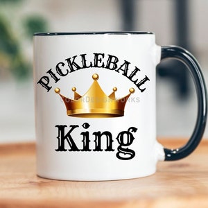 May include: White ceramic coffee mug with a black rim and handle. The mug has a black and gold design that says "Pickleball King" with a gold crown.