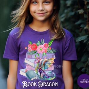 May include: A purple t-shirt featuring a watercolor design of a dragon, books, and flowers. The text "Book Dragon" is below the image. A purple circle with the words "Team Purple" is in the lower right corner.