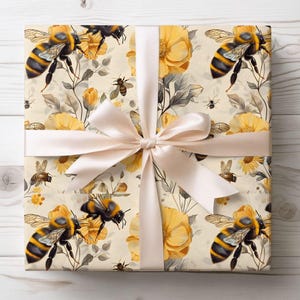 May include: Gift wrap featuring a repeating pattern of yellow and black bees, yellow flowers, and gray leaves on a cream background. A cream-colored satin ribbon is tied in a bow around the wrapped package.