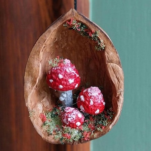 Könnte beinhalten: Ein kleines, dekoratives Ornament aus einer Walnussschale mit drei roten und weißen Pilzen darin. Die Pilze sind mit weißen Punkten bemalt und haben eine schneeweiße Beschichtung auf der Oberseite.