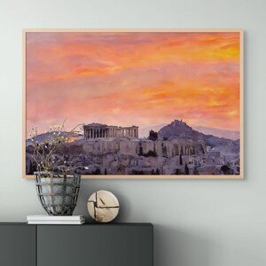 May include: Framed print of the Acropolis in Athens, Greece, under a vibrant sunset sky. The artwork features the ancient structures in shades of gray and beige, set against a backdrop of orange and pink clouds. The print is in a light-colored wooden frame.