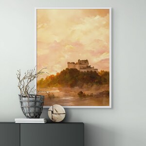 May include: A framed print of a watercolor painting depicting a castle on a hilltop with a misty river in the foreground. The sky is a soft orange and yellow, with fluffy white clouds.