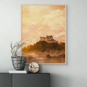 May include: A framed watercolor painting of a castle on a hilltop with a misty river in the foreground. The sky is a soft orange and pink.