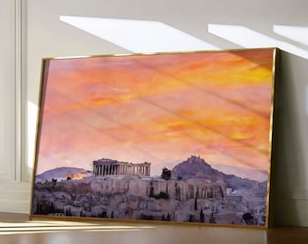May include: A framed print of the Acropolis in Athens, Greece, featuring a vibrant sunset sky in shades of orange and yellow. The print has a gold-colored frame and is leaning against a wall.