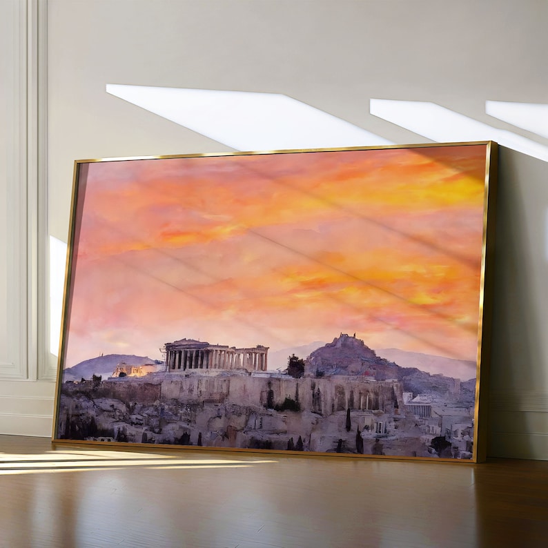 May include: A framed print of the Acropolis in Athens, Greece, featuring a vibrant sunset sky in shades of orange and yellow. The print has a gold-colored frame and is leaning against a wall.