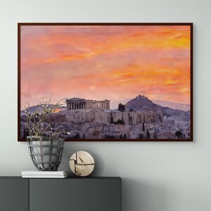 May include: Framed print of the Acropolis in Athens, Greece, under a vibrant sunset sky. The artwork features the ancient structures in shades of gray and beige, set against a backdrop of orange and pink clouds. The frame is a dark brown.