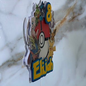 Puede incluir: Un adorno para pastel colorido con un grupo de personajes de Pok&eacute;mon, incluyendo Pikachu, Bulbasaur y Charmander, que rodean una Pok&eacute; Ball. El adorno tambi&eacute;n incluye el n&uacute;mero 8 y el nombre "Elijah".