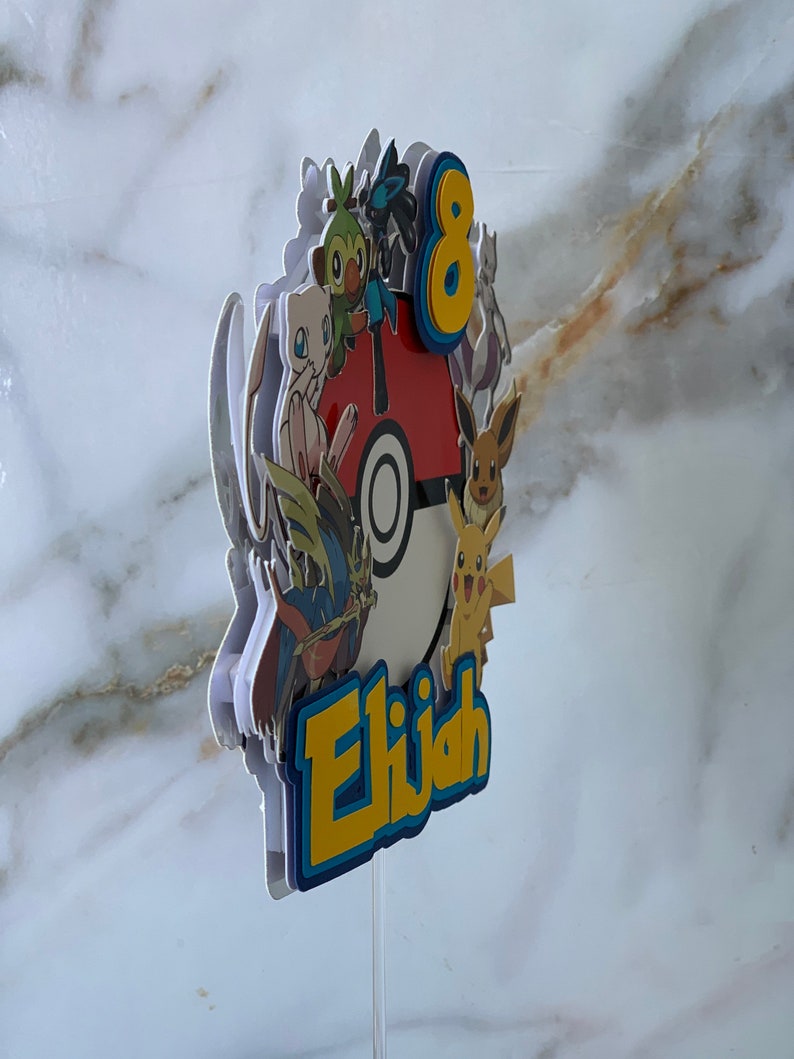Puede incluir: Un adorno para pastel colorido con un grupo de personajes de Pok&eacute;mon, incluyendo Pikachu, Bulbasaur y Charmander, que rodean una Pok&eacute; Ball. El adorno tambi&eacute;n incluye el n&uacute;mero 8 y el nombre "Elijah".