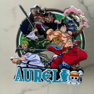 以下が含まれることがあります： ワンピースのアニメケーキトッパー。緑、白、赤の3人のキャラクターが描かれています。キャラクターはルフィ、ゾロ、シャンクスです。トッパーには「AUREL」の名前とワンピースのドクロとクロスボーンのロゴも含まれています。