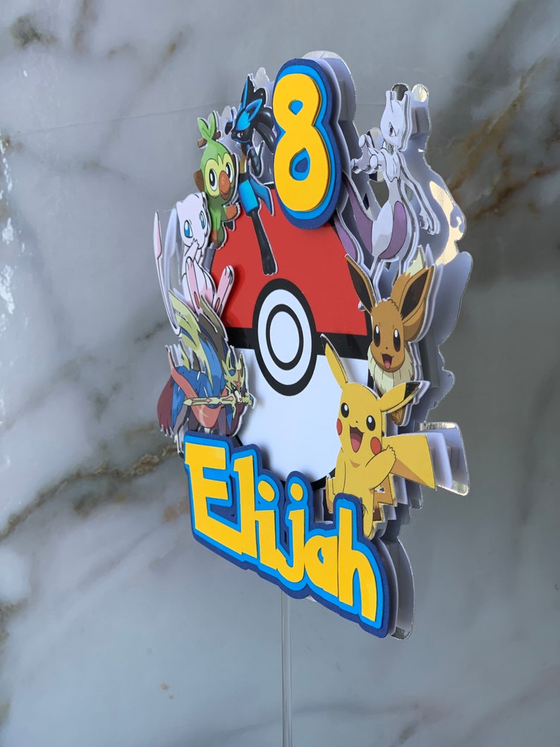 Puede incluir: Un adorno para pastel de Pok&eacute;mon amarillo, azul y rojo con el n&uacute;mero 8 y el nombre Elijah. El adorno presenta varios personajes de Pok&eacute;mon, incluidos Pikachu, Eevee y Grookey.