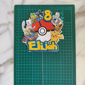 Puede incluir: Un adorno para pastel con una Pok&eacute; Ball roja y blanca con el n&uacute;mero 8 y el nombre Elijah en letras amarillas. El adorno est&aacute; rodeado de varios personajes de Pok&eacute;mon, incluyendo Pikachu, Bulbasaur, Charmander, Squirtle y otros.