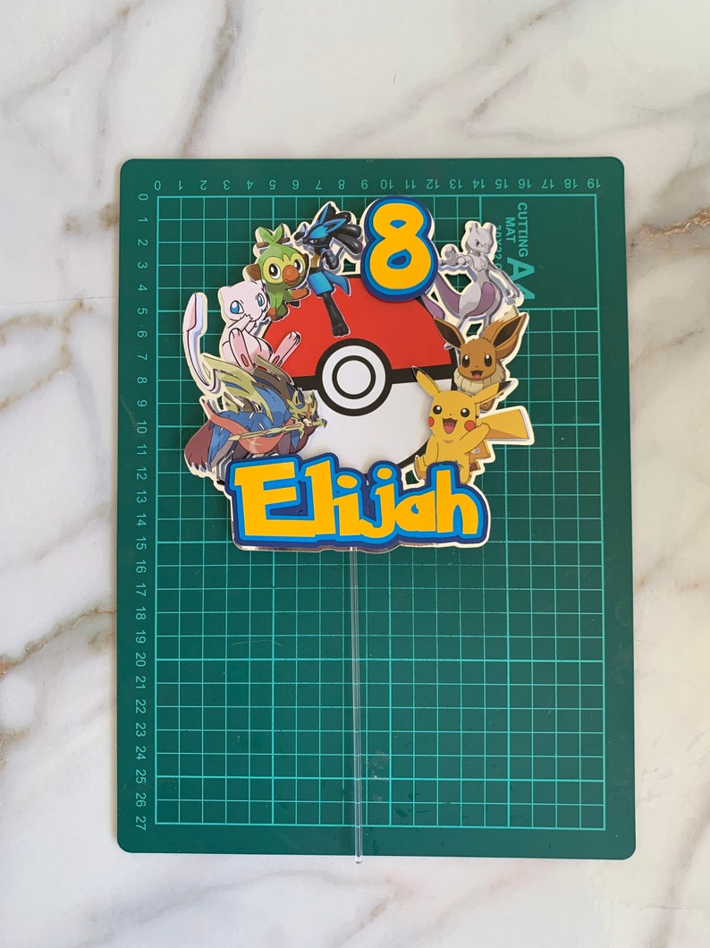 Puede incluir: Un adorno para pastel con una Pok&eacute; Ball roja y blanca con el n&uacute;mero 8 y el nombre Elijah en letras amarillas. El adorno est&aacute; rodeado de varios personajes de Pok&eacute;mon, incluyendo Pikachu, Bulbasaur, Charmander, Squirtle y otros.