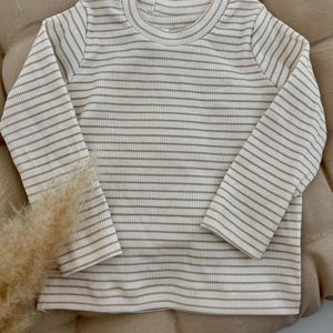 Könnte beinhalten: Ein langärmliges Shirt aus geripptem Strick in cremefarbenen und braunen horizontalen Streifen. Das Shirt hat einen Rundhalsausschnitt und ist aus einem weichen, bequemen Material gefertigt. Die neutrale Farbpalette macht es vielseitig.
