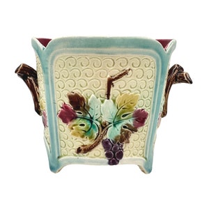 May include: A square ceramic planter with a light blue glaze and a raised design of grape leaves and grapes in shades of green, brown, and purple. The planter has two brown handles on the sides.