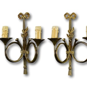 May include: A pair of gold-toned wall sconces with a French horn design. Each sconce has two candle-style light bulbs.