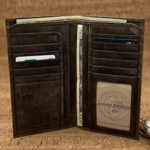May include: Open brown leather wallet with multiple card slots, cash compartments, and a clear ID window. The wallet is open, revealing cash and cards. The wallet has a distressed finish and the text "Emerald Engraving Co."