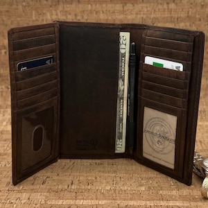 Puede incluir: Cartera de cuero marrón abierta con múltiples ranuras para tarjetas, una ventana de identificación transparente y un bolsillo que contiene un billete de veinte dólares y un bolígrafo negro. La cartera está abierta, mostrando su interior.